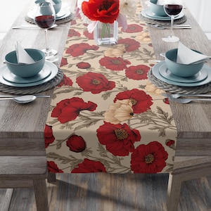 May include: A beige table runner featuring a floral pattern of red and cream-colored poppies with green stems and leaves. The runner is set on a wooden table, with place settings and a vase of flowers.