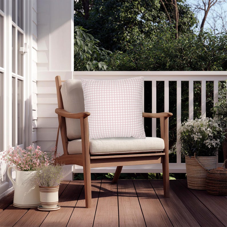 May include: A wooden outdoor chair with a light beige cushion and a pink and white gingham patterned pillow. The chair is on a wooden deck, with white railing and greenery in the background. Decorative flower pots are on the deck.