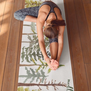 May include: A yoga mat with a watercolor design of trees in shades of green and brown. The mat has the name "nicolle" printed on the bottom right corner. A person is doing a yoga pose on the mat.