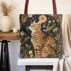 Puede incluir: Bolso tote con una ilustración detallada de un gato atigrado en un diseño floral. El gato tiene rayas marrones y naranjas, mirando hacia arriba. El fondo es verde oscuro con flores amarillas y naranjas. El bolso tiene correas burdeos.