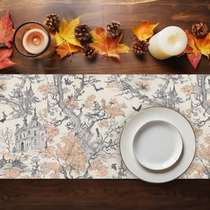 Puede incluir: Una mesa decorada para Halloween con un camino de mesa crema con un diseño de árbol y casa embrujada en negro y melocotón. La mesa está decorada con hojas de otoño, piñas, una vela en un portavelas de cristal y un plato blanco con borde dorado.