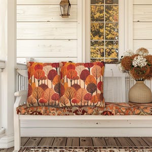 May include: Two orange, red, and brown throw pillows with a forest pattern on a white bench with a floral patterned cushion. The bench is on a porch with a white wooden wall and a window with fall foliage visible through the glass.