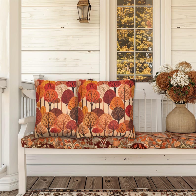 May include: Two orange, red, and brown throw pillows with a forest pattern on a white bench with a floral patterned cushion. The bench is on a porch with a white wooden wall and a window with fall foliage visible through the glass.