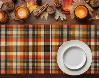 May include: Overhead view of a fall-themed table setting. A plaid table runner in orange, brown, and cream colors is topped with white plates. Lit candles, fall leaves, pine cones, and cinnamon sticks decorate the wooden surface.