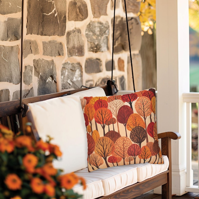 May include: A white cushion with a fall-themed pillow featuring a pattern of orange, red, and brown trees on a cream background. The pillow is on a wooden porch swing.