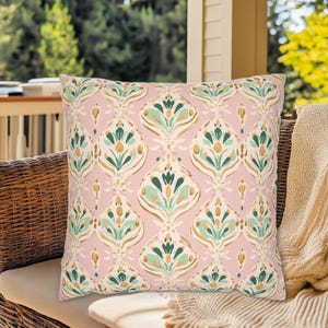 May include: Square decorative pillow with a repeating floral pattern in shades of pink, green, gold, and cream. The pillow is placed on a wicker chair with a neutral-colored blanket draped over it. The background shows a porch and trees.