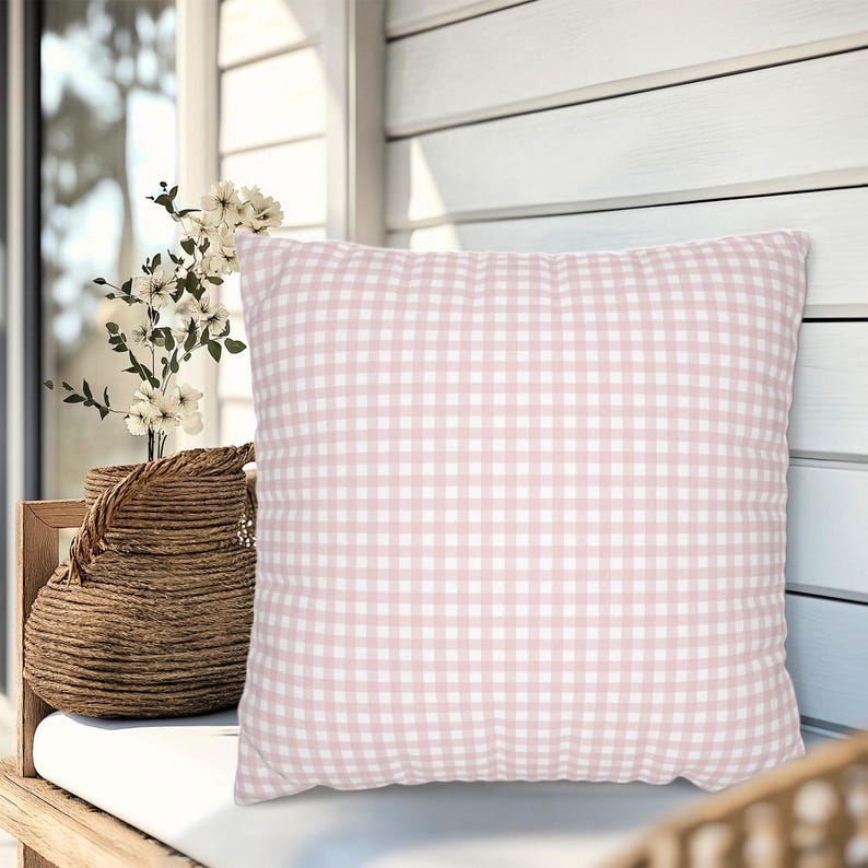 May include: A square decorative pillow with a pink and white gingham pattern. The pillow is placed on a white cushion, with a woven basket and flowers in the background. The pillow is a soft, inviting accent for any room.