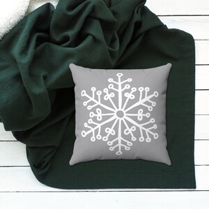 Puede incluir: Una almohada gris con un dise&ntilde;o de copo de nieve blanco sobre una manta de color verde oscuro.