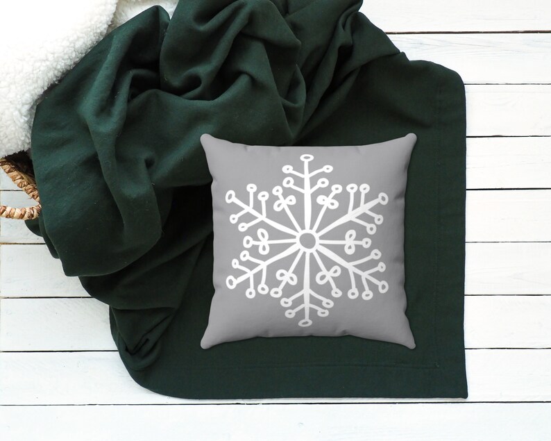 Puede incluir: Una almohada gris con un dise&ntilde;o de copo de nieve blanco sobre una manta de color verde oscuro.