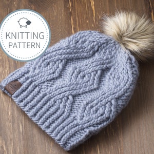 May include: A light blue knitted hat with a cable knit design and a faux fur pom-pom. The hat has a small brown leather tag. The image also includes a circular graphic that says "Knitting Pattern".