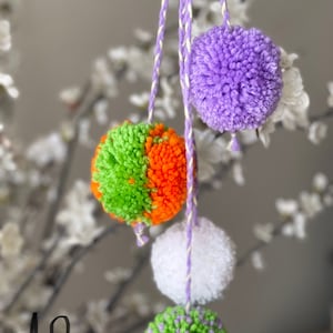 May include: A set of four colourful pom-poms hanging from strings. The pom-poms are green and orange, white, purple, and green and purple.