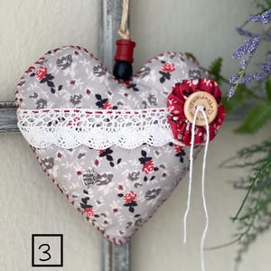 Könnte beinhalten: Ein graues und rotes herzförmiges Kissen mit einer weißen Spitzenborte und einem kleinen silbernen Anhänger mit der Aufschrift "Made with Love". Das Kissen hat einen roten Knopf mit der Aufschrift "Handmade" darauf.