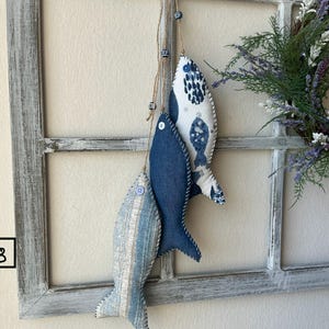 May include: Three fabric fish decorations in shades of blue and white hang from twine against a distressed white window frame. One fish is solid blue, another has a blue and white pattern, and the third has a striped pattern. A lavender wreath is in the background.