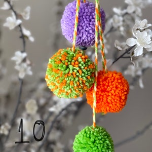 May include: Four yarn pom-poms in purple, green and orange, and solid green, hanging from braided orange and green strings. The number 10 is visible on the left side. The pom-poms are made of soft, fuzzy yarn fibers in bright, distinct colors.