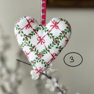 May include: White heart-shaped fabric ornament with a red and green holly and bow pattern. The ornament is hanging from a red and white ribbon.