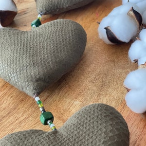 May include: Three green felt heart-shaped cushions with a string of green, white and yellow beads connecting them. The hearts are on a wooden surface with white cotton balls in the background.