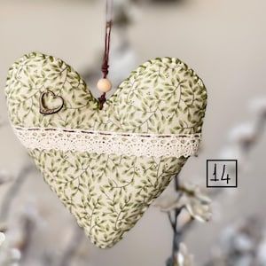 Puede incluir: Un adorno de tela en forma de corazón verde y blanco con un borde de encaje y un pequeño dije de corazón de metal. El adorno está colgado de una cuerda marrón con una cuenta de madera. El número 14 es visible en la esquina inferior derecha de la imagen.