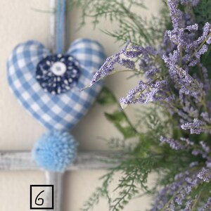 May include: A blue and white gingham heart-shaped ornament with a blue pom-pom and a floral embellishment. The ornament hangs from a blue ribbon. Lavender-colored flowers and greenery are in the background.