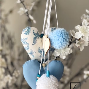 May include: A hanging decoration featuring three blue and white heart-shaped fabric ornaments. The hearts are adorned with a floral pattern and have blue and white pom-poms attached. The decoration is suspended from a white string.