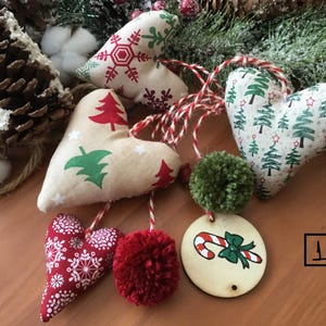 Könnte beinhalten: Weihnachtsschmuck in Herzform und ein runder Holzanhänger. Die Herzen zeigen festliche Muster wie Schneeflocken, Weihnachtsbäume und Sterne. Rote und grüne Pompons begleiten die Ornamente.