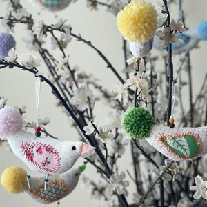 May include: A white blossom branch with handmade fabric bird ornaments. The birds have pom-poms for heads and are decorated with colorful embroidery.