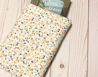 May include: A floral book sleeve with a book titled "orphan train" peeking out. The sleeve is cream-colored with a pattern of small peach, yellow, and navy blue flowers. The book is a #1 New York Times bestseller.