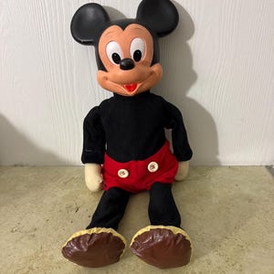 May include: A vintage Mickey Mouse plush doll. The doll has a black body, red shorts with white buttons, and large brown shoes. Mickey's face is tan with black eyes, a red mouth, and large black ears. The doll is sitting against a white wall.