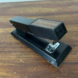 May include: A black stapler with a wood-grain top and a silver staple remover. The stapler is made by the brand "BATES".