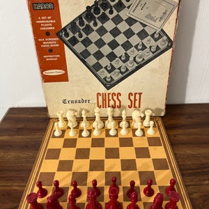 Puede incluir: Un juego de ajedrez vintage Baron Crusader con un tablero marrón y beige y piezas de ajedrez de plástico rojo y blanco. La caja está etiquetada como "Crusader Chess Set" y presenta un tablero de ajedrez blanco y negro con piezas sobre él.