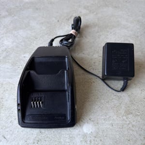 May include: A black Motorola OverNight charging cradle with a recessed slot for a device, featuring four metal contact pins at the base. The unit includes an attached black power cord connected to a rectangular black wall plug adapter.