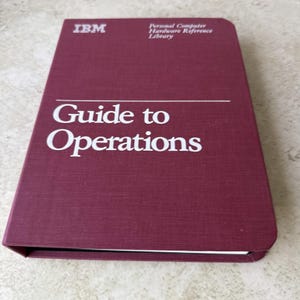 May include: A maroon IBM Personal Computer Hardware Reference Library book titled Guide to Operations. The cover features white text and a horizontal line, with a textured fabric-like finish and a rounded corner on the top right side.