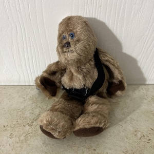 Op de afbeelding: Een pluche speelgoed van een Wookiee-personage, met bruine vacht, blauwe ogen en een zwarte bandolier. Het speelgoed zit op een lichtgekleurd oppervlak en heeft donkerbruine accenten op handen en voeten.