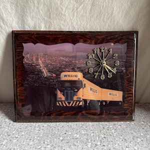 May include: A rectangular wall clock featuring a cityscape backdrop and a yellow Willig semi-truck. The clock face has gold-coloured hands and numerals. The frame has a dark wood-grain finish.