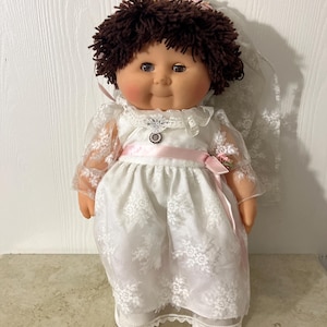 May include: A vintage doll with brown yarn hair, dressed in a white lace wedding dress with a pink ribbon sash. The doll has rosy cheeks, blue eyes, and a lace veil. A small pendant is at the neckline.