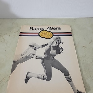 Può includere: Copertina di un programma di calcio vintage con un'immagine in bianco e nero di un giocatore che corre con la palla. La copertina presenta la scritta "Rams vs 49ers" con un emblema dorato e strisce rosse, bianche e blu.