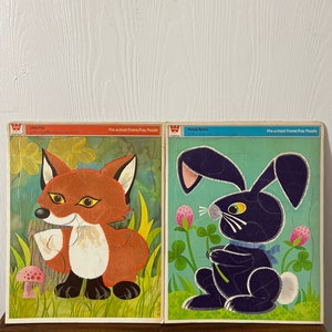 Può includere: Due puzzle vintage con cornice e vassoio per la scuola materna. Uno presenta una volpe dei cartoni animati con pelliccia arancione, dettagli bianchi e un fungo rosa. L'altro puzzle raffigura un coniglietto nero con un fiocco blu, circondato da trifogli. I puzzle sono intitolati "Little Fox" e "Purple Bunny".
