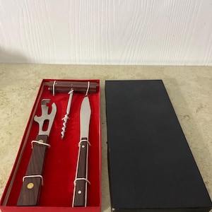 Puede incluir: Un juego de abridor de vino en una caja roja con tapa negra. El juego incluye un abridor de botellas, un sacacorchos y un cuchillo, todos con mangos de madera oscura y componentes de metal plateado. La caja es rectangular.