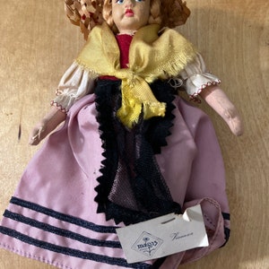 May include: A vintage doll dressed in a straw hat, red top, and pink skirt with black trim. The doll has blonde hair and red shoes. A tag reads "Vienzia".
