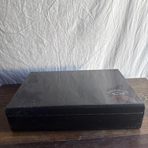 Puede incluir: Una caja rectangular negra con un aspecto ligeramente desgastado. La caja tiene un logotipo en la esquina superior derecha con el texto "PAUL & SHARK". La caja está sobre una superficie de madera.