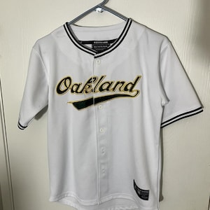 May include: White baseball jersey with the word "Oakland" in gold and green script across the chest. The jersey has black trim on the collar and sleeves, and a button-up front. The word "RENEGADE" is printed on the collar.