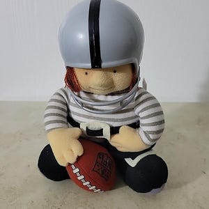 May include: A soft toy of a football player wearing a grey helmet with a black stripe and a grey and white striped top. The doll holds a brown football with white laces and the NFL logo.