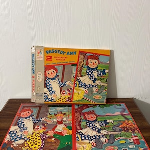 Può includere: Una scatola vintage di Raggedy Ann 2 Elementary Puzzles con grandi pezzi che si incastrano. La scatola presenta un'illustrazione colorata di Raggedy Ann con un vestito a quadri rosso e bianco e un fiocco rosso. La scatola è etichettata "MB Easy To Fun" e "Milton Bradley Company".