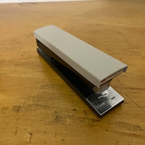 Puede incluir: Una grapadora negra y blanca con la parte superior blanca y la base negra. La grapadora tiene un quita grapas de metal plateado.