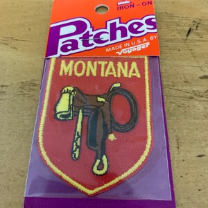 May include: Iron-on patch featuring a brown saddle and reins on a red background with a yellow border. The text "MONTANA" is printed in yellow on a yellow banner above the saddle. The patch is packaged in a clear plastic bag with a purple and orange header that reads "Patches" and "Made in U.S.A. by Voyager".
