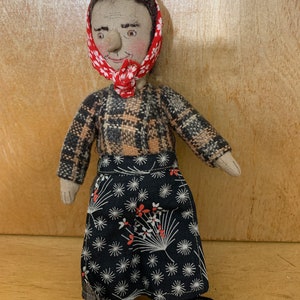 May include: Handmade doll with a fabric face, wearing a plaid jacket, a black skirt with white and red floral designs, and a red floral headscarf. The doll has black shoes and brown hair.