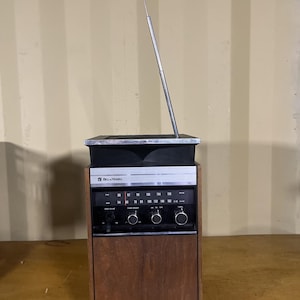 Könnte beinhalten: Vintage Bell & Howell Radio mit Holzgehäuse und silberner Antenne. Das Radio hat ein schwarzes Bedienfeld mit Drehreglern und eine Frequenzanzeige. Das Design ist retro, mit einer klassischen Ästhetik der Mitte des Jahrhunderts.