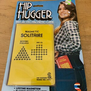 Puede incluir: Un juego magnético "Hip Hugger" vintage. El embalaje presenta un juego "Magnetic Solitaire" amarillo, un juego "Magnetic Checkers" rojo y una mujer con una camisa a cuadros y jeans. El embalaje también incluye el texto "Take Along Magnetic Game".