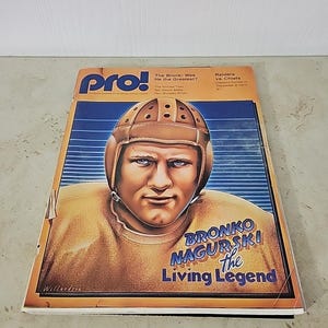 May include: A vintage "pro!" magazine cover featuring a portrait of Bronko Nagurski in a football helmet and jersey. The cover has text that reads "Bronko Nagurski the Living Legend" and "Raiders vs. Chiefs". The magazine is on a white surface.