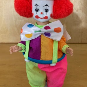 May include: A vintage clown doll with a white face, red hair, and a polka-dot hat. The doll wears a colorful outfit with a large bow tie and suspenders. The clown's outfit includes purple, orange, pink, and green sections.