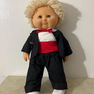 Könnte beinhalten: Eine Vintage-Puppe mit blonden, garnartigen Haaren, die einen schwarzen Smoking, eine Hose und weiße Schuhe trägt. Die Puppe hat eine rote Fliege und einen Kummerbund. Die Puppe steht auf einer hellen Oberfläche.